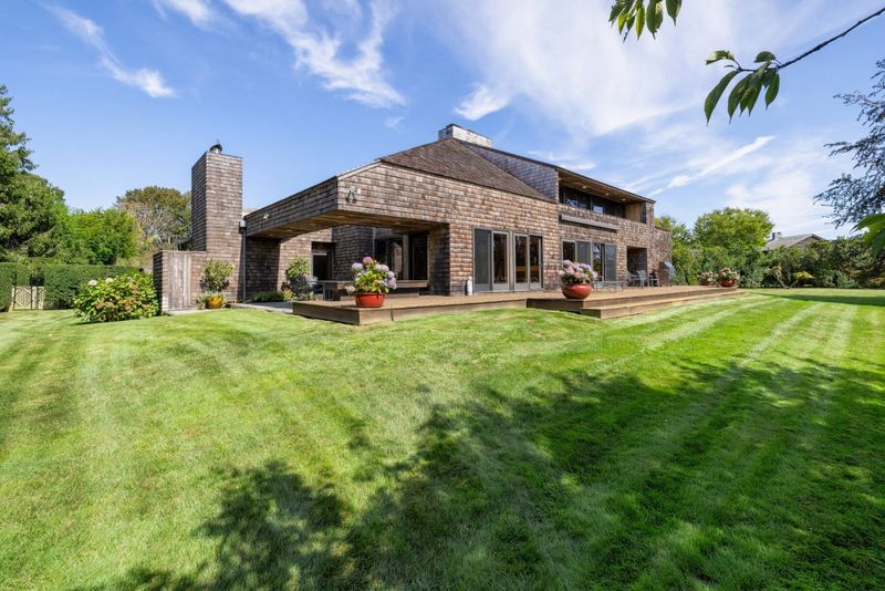 The rear facade from the striped lawn: the pyramidal roof, shingled walls and a wall of glass under deep overhangs, with mature trees at the edges. at 95 Jobs Lane, Bridgehampton — photo 3 of 12