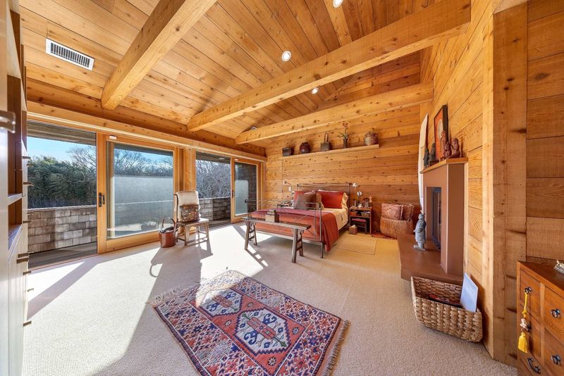 The primary suite under the peaked roof: exposed beams, a rug-covered floor, a wall of glass onto a terrace and a fireplace. at 95 Jobs Lane, Bridgehampton — photo 10 of 12